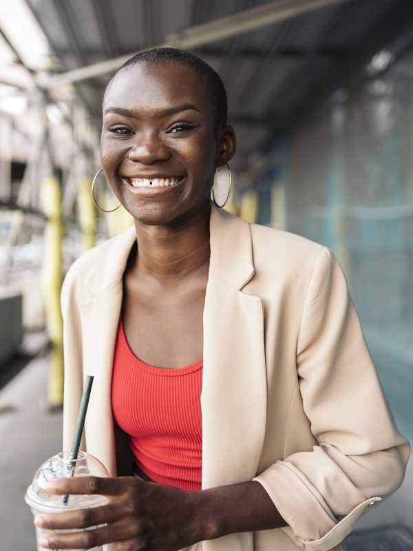 Vertical image of a cheerful elegant young african american woman holding a milkshake standing under scaffolding in a construction site looking at camera.